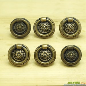 May include: Six antique brass cabinet knobs with a ring pull and a decorative floral design. The knobs are arranged in two rows of three on a wooden surface.