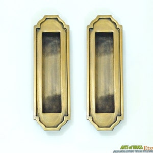May include: Two antique brass cabinet door pulls with a rectangular shape and an ornate design. The pulls have a slightly aged finish.
