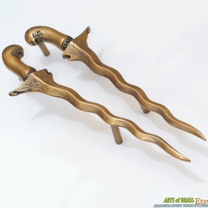 Op de afbeelding: Een paar sierlijke messing deurgrepen in de vorm van dolken met ingewikkelde ontwerpen. De grepen zijn gebogen en hebben een puntige punt.