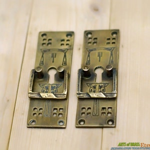 May include: Two antique brass door handle plates with keyhole and decorative handles. Each plate features an Art Nouveau design with geometric and floral motifs. The handles have a rectangular shape with rounded edges and two cylindrical posts.