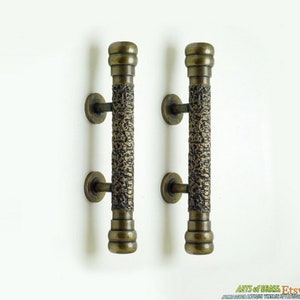 Może przedstawiać: Dwa antyczne mosiężne klamki drzwiowe z fakturowanym wykończeniem. Klamki są długie i cylindryczne z zaokrąglonym górnym i dolnym końcem.