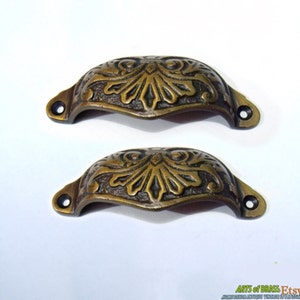 Può includere: Due tiretti per cassetti in ottone antico con un design floreale ornato. I tiretti sono lunghi 7,6 cm e hanno una forma curva.