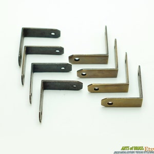 Puede incluir: Ocho soportes de esquina de latón antiguo, algunos con una pátina más oscura, dispuestos sobre una superficie blanca. Los soportes tienen forma de L y tienen un agujero en cada extremo.