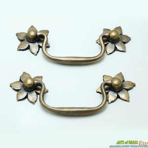May include: Two antique brass drawer pulls with a floral design. Each pull has a rounded, slightly curved handle and a decorative flower-shaped knob.