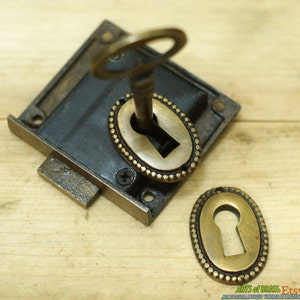 May include: Antique brass keyhole escutcheon with a key in the keyhole. The escutcheon has a decorative beaded border.