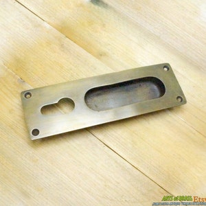 May include: A brass door pull with a rectangular shape and a long oval cutout. The pull has two small holes on either side for mounting.