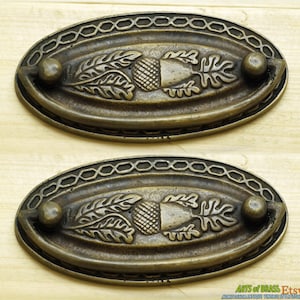 May include: Two antique brass cabinet pulls featuring an acorn and leaf design. The pulls are oval shaped with a decorative border and a small knob on each side.