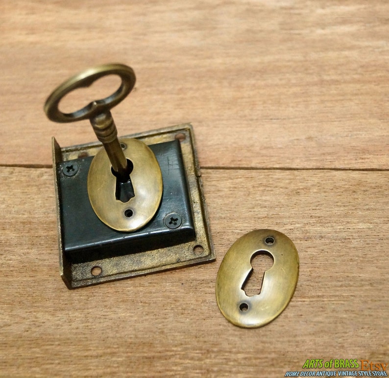 May include: A brass keyhole cover with a key inserted into the keyhole. The cover is attached to a square brass plate with four screws. The keyhole cover is oval shaped and has a decorative edge. The key is ornate and has a decorative handle.