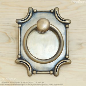 May include: A square brass door pull with a ring handle. The pull has a decorative design with rounded edges and a textured finish.