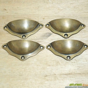 May include: Four antique brass cup pulls with a scalloped edge and three holes. The pulls are arranged in two rows of two on a wooden surface.