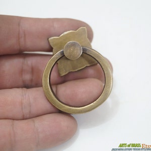 Puede incluir: Un tirador de anillo de lat&oacute;n con un dise&ntilde;o decorativo en la parte posterior. El tirador de anillo tiene un di&aacute;metro de aproximadamente 5 cm.