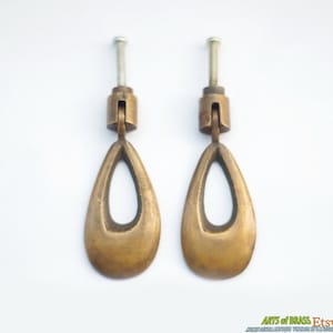 Può includere: Due maniglie per porte di armadio in ottone a forma di lacrima con un ovale tagliato al centro. Le maniglie hanno una finitura spazzolata e sono fissate a una vite.