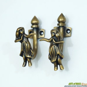 May include: Two brass hooks with a decorative design of a woman in a flowing skirt. The hooks are mounted on a white wall.