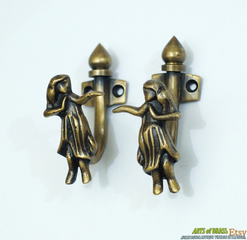 May include: Two brass hooks with a decorative design of a woman in a flowing skirt. The hooks are mounted on a white wall.