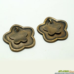 Może przedstawiać: Dwa zawieszki w kształcie kwiatu wykonane z mosiądzu, z mniejszym kwiatem wewnątrz każdego. Zawieszki mają pętelkę na górze do zawieszenia.