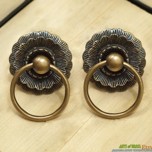 May include: Two antique brass drawer pulls with a floral design and a ring handle. The pulls are ornate and have a distressed finish.