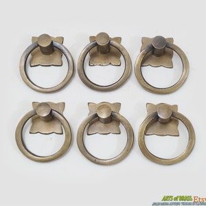 May include: Six antique brass ring pulls with a square base. The pulls have a smooth, aged finish and are perfect for adding a touch of vintage charm to furniture or cabinets.