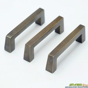 May include: Three antique brass cabinet pulls with a rectangular shape and a slightly curved handle. The pulls have a distressed finish and a rustic look.