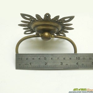 May include: A brass drawer pull with a decorative, ornate design. The pull is 2 inches wide and has a ring handle.