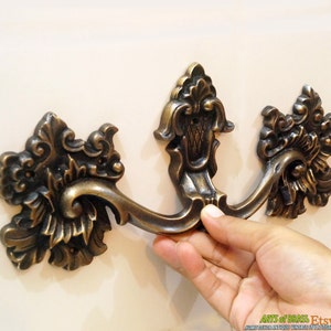 May include: Ornate brass drawer pull with a decorative floral design. The pull has a curved shape and a centre section with a raised design. The pull is mounted on a white surface.