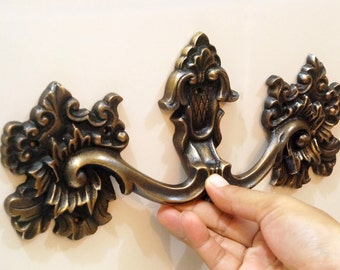 May include: Ornate brass drawer pull with a decorative floral design. The pull has a curved shape and a center section with a raised design. The pull is mounted on a white surface.