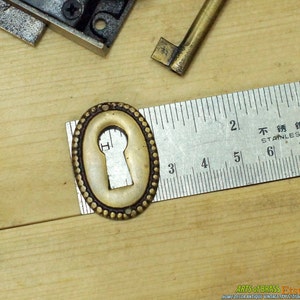 May include: Brass keyhole cover with a decorative beaded edge. The cover is oval shaped and measures approximately 1 1/2 inches in length.
