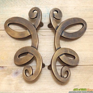 May include: A pair of antique brass drawer pulls with a decorative, swirling design. The pulls are in the shape of a stylised butterfly.
