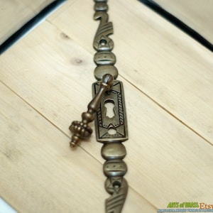 May include: Antique brass drawer pull with a keyhole and decorative details. The pull is attached to a long, ornate brass plate with a repeating pattern.