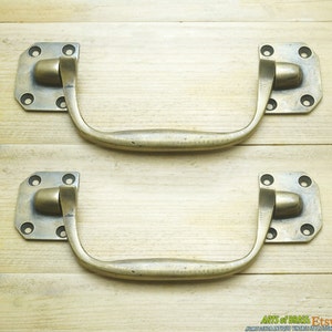 May include: Two antique brass cabinet handles with a curved design and a square backplate. Each handle has four decorative holes on the backplate.