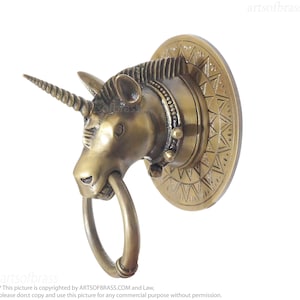 May include: A brass unicorn door knocker. The knocker features a detailed unicorn head with a spiraled horn and a ring in its mouth. The round backplate has a geometric pattern. The text "artsofbrass" is visible.