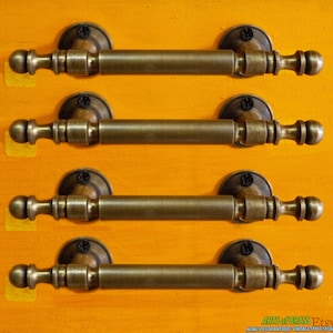 May include: Four antique brass drawer pulls with rounded ends and a cylindrical center. The pulls are arranged in a row on a yellow background.