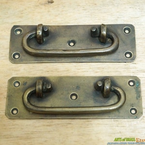 May include: Two antique brass drawer pulls with a rounded rectangular base and a curved handle. The pulls have a worn, aged finish.
