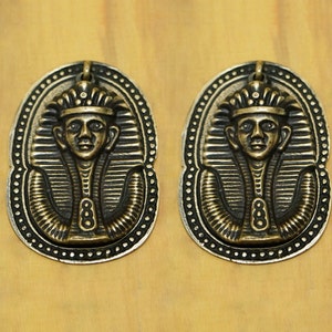 May include: Two gold-toned metal pendants featuring an Egyptian pharaoh design. Each pendant has a detailed depiction of a pharaoh's head with a crown and a patterned collar.