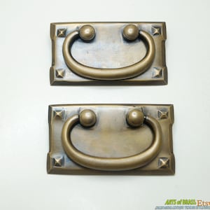 Könnte beinhalten: Zwei antike Messing-Schubladengriffe mit rechteckiger Basis und abgerundetem, gewölbtem Griff. Die Griffe haben eine patinierte Oberfläche und ein dekoratives Design an den Rändern.