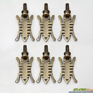 Puede incluir: Seis pomos de armario de latón antiguo con un diseño geométrico. Los pomos tienen la forma de un insecto estilizado con una parte inferior puntiaguda y un bucle en la parte superior para sujetarlo a una puerta de armario.