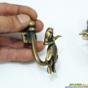 May include: Two antique brass hooks with a decorative design of a woman in a dress. The hooks are attached to a wall and are ready to hang items.