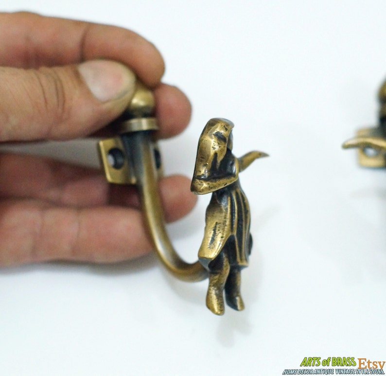 May include: Two antique brass hooks with a decorative design of a woman in a dress. The hooks are attached to a wall and are ready to hang items.
