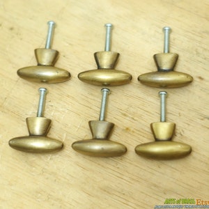 May include: Six oval-shaped brass cabinet knobs with a raised center and a screw. The knobs are a light gold color and have a vintage look.