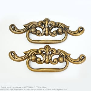 May include: Two ornate gold-toned metal drawer pulls with a fleur-de-lis design. Each pull has a rectangular opening and a decorative scrollwork design.