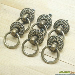 May include: Six antique brass drawer pulls with ornate floral designs and ring handles. The pulls are arranged in a semi-circle on a wooden surface.