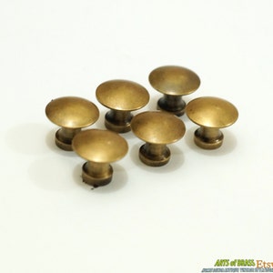 May include: Six antique brass mushroom-shaped cabinet knobs. Each knob has a rounded top and a short, cylindrical base. The knobs are arranged in a cluster on a white background. The knobs have a warm, aged brass finish.