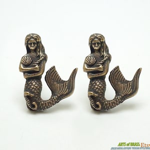 Peut inclure: Une paire de tirettes de tiroir en laiton antique en forme de sirène. Chaque tirette représente une sirène tenant une coquille dans une main et une queue avec des écailles. Les sirènes sont très détaillées et en bon état.