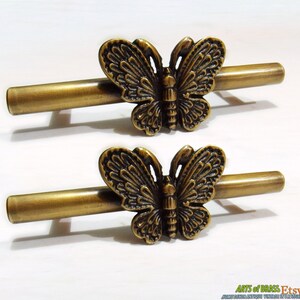 Op de afbeelding: Twee antieke messing vlindergrepen voor lades. Elke greep heeft een gedetailleerd vlinderontwerp met ingewikkelde vleugels en een getextureerd lichaam. De grepen zijn bevestigd aan een gladde, cilindrische stang.