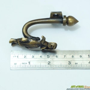 May include: Brass curtain tieback with a figure of a person holding a drapery hook. The tieback measures approximately 5 inches long.