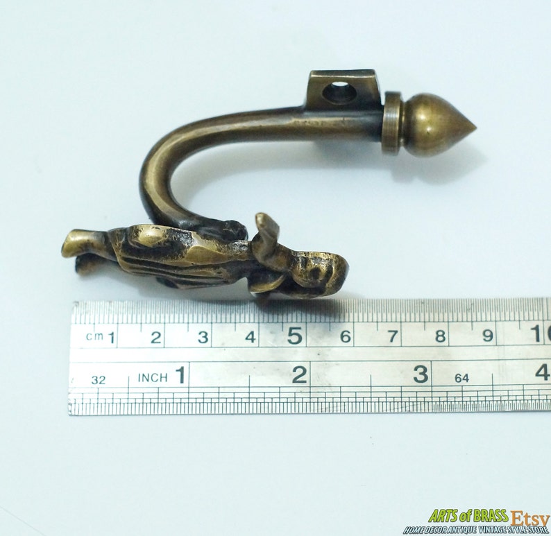 May include: Brass curtain tieback with a figure of a person holding a drapery hook. The tieback measures approximately 5 inches long.