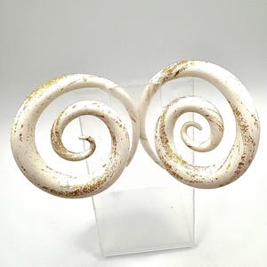 May include: A pair of white spiral ear gauges with gold accents. The gauges are made of a smooth, polished material and have a subtle distressed look.