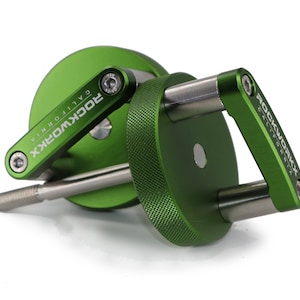 May include: A green metal tool with the text "ROCKWORXX CALIFORNIA" on the side. The tool has a circular base and a handle, with a silver-colored rod extending from the center. The handle and base have a textured pattern.