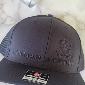 May include: Black trucker hat with a black embroidered skull and crossbones design and the text "Lineman Attitude". The hat is made by Richardson and has a black mesh back.