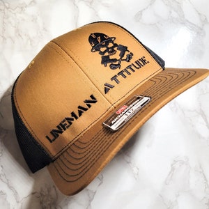 May include: A brown and black baseball cap with a black embroidered skull wearing a hard hat and holding crossed wrenches. The text "LINEMAN ATTITUDE" is embroidered on the cap.