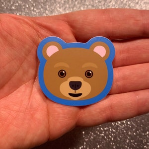 May include: A brown bear face sticker with a blue outline. The bear has pink ears, brown fur, and a friendly smile.